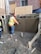 A construction worker wearing a hard hat and safety vest is applying a finish to a freshly laid concrete wall. The area is surrounded by muddy ground, and wooden and metal frameworks are visible. The background includes a brick wall and wooden boards.