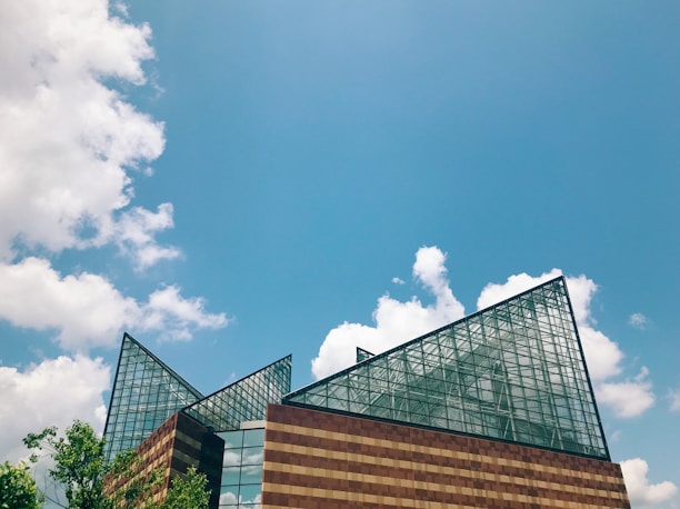 A modern building with large, triangular glass panels against a backdrop of a bright blue sky with scattered white clouds. The building features a mix of transparent and reflective surfaces, with an angular architectural style. Green foliage is visible in the foreground, adding a natural element to the urban landscape.