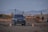 A black SUV is driving on a rocky terrain with orange traffic cones placed around it. The background features distant mountains under a slightly cloudy sky and some greenery. There appears to be a structure resembling a minaret in the distance.