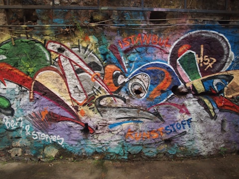 Colorful graffiti covers a textured stone wall, featuring a mix of abstract shapes and vibrant patterns in shades of green, red, blue, and orange. The words 'Istanbul' and 'KUNSTSTOFF' are visible among the designs.