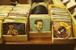 Stacks of vintage vinyl records are arranged in cardboard boxes on a wooden surface. The records feature colorful and artistic album covers with illustrations and photographs of artists. The collection includes various records from different music genres, showcasing a retro and nostalgic vibe.