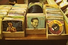 Stacks of vintage vinyl records are arranged in cardboard boxes on a wooden surface. The records feature colorful and artistic album covers with illustrations and photographs of artists. The collection includes various records from different music genres, showcasing a retro and nostalgic vibe.