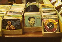 Stacks of vintage vinyl records are arranged in cardboard boxes on a wooden surface. The records feature colorful and artistic album covers with illustrations and photographs of artists. The collection includes various records from different music genres, showcasing a retro and nostalgic vibe.