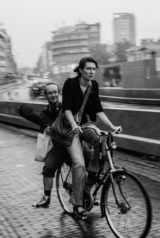 A black-and-white image of two individuals riding a bicycle amidst urban surroundings. The person in front pedals with a determined expression, while the one at the back appears relaxed, holding a shopping bag. Buildings and vehicles are visible in the blurred background, suggesting motion and a bustling city environment.