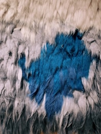 A textured surface with long, flowing strands of fur-like material in various colors. The top section features off-white hues that blend into gray, and further down there is a prominent deep blue area. The strands appear soft and shaggy, giving a sense of warmth and comfort.