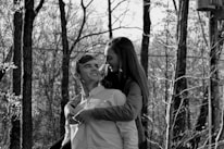 A black and white photo depicting a young couple in a forested area, where the woman is playfully embracing the man from behind. Both appear to be smiling and looking at each other, set against a background of bare trees and natural scenery.
