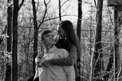 A black and white photo depicting a young couple in a forested area, where the woman is playfully embracing the man from behind. Both appear to be smiling and looking at each other, set against a background of bare trees and natural scenery.