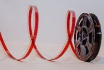 A film reel with a length of film unwound and extended in a looping pattern. The film is red and perforated along the edges and the reel is dark gray or black.