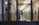 A long corridor in a prison with multiple closed cell doors lining both sides. The lighting is dim and fluorescent, creating a stark and sterile atmosphere. The perspective is through metal bars, suggesting confinement.