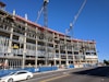 A large multi-story building under construction with visible scaffolding and cranes. The structure appears to be made of concrete and steel, with an open frame on several floors. A white car is driving on the road in front of the construction site, and there are safety barriers and signage visible along the sidewalk.