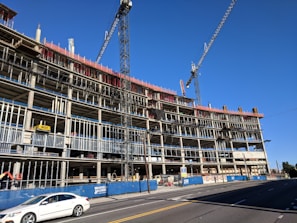 A large multi-story building under construction with visible scaffolding and cranes. The structure appears to be made of concrete and steel, with an open frame on several floors. A white car is driving on the road in front of the construction site, and there are safety barriers and signage visible along the sidewalk.