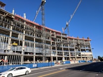 A large multi-story building under construction with visible scaffolding and cranes. The structure appears to be made of concrete and steel, with an open frame on several floors. A white car is driving on the road in front of the construction site, and there are safety barriers and signage visible along the sidewalk.