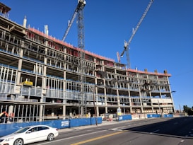 A large multi-story building under construction with visible scaffolding and cranes. The structure appears to be made of concrete and steel, with an open frame on several floors. A white car is driving on the road in front of the construction site, and there are safety barriers and signage visible along the sidewalk.
