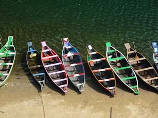A row of colorful wooden boats neatly aligned on a sandy shore beside a body of water with a greenish hue. Each boat features distinct painted patterns, including stripes and shapes, in various colors such as green, blue, red, and white. The boats are positioned with their bows facing the water, and some have paddles or other items inside.