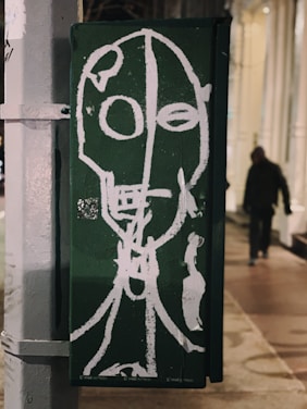 A piece of street art is created on a green background featuring an abstract white outline of a face with circular eyes and an elongated neck. The style is raw and expressive, painted on an urban structure possibly a utility box on a sidewalk. In the background, a blurred figure walks by, adding a sense of street life to the scene.