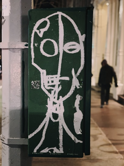 A piece of street art is created on a green background featuring an abstract white outline of a face with circular eyes and an elongated neck. The style is raw and expressive, painted on an urban structure possibly a utility box on a sidewalk. In the background, a blurred figure walks by, adding a sense of street life to the scene.