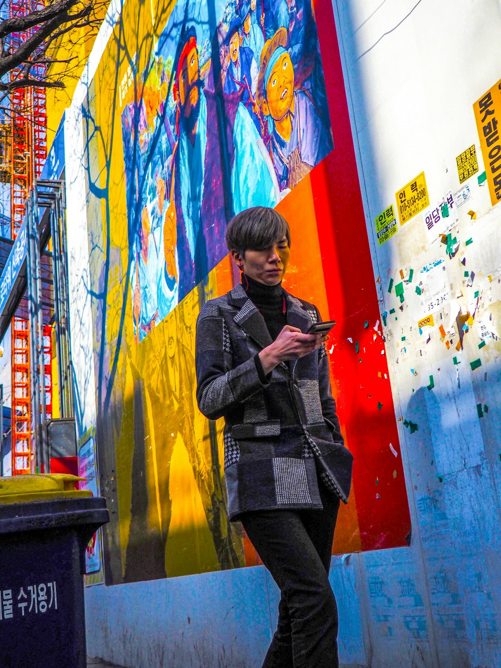 person in gray coat holding smartphone while walking near graffiti wall