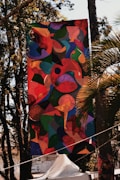 A large, colorful abstract banner is hanging outdoors among trees. The artwork features a mixture of organic shapes in vibrant colors, including reds, greens, blues, and yellows. Sunlight filters through the trees, casting shadows and highlights on the banner. Below the banner, there is a white tent and some signage visible in the background.