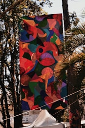 A large, colorful abstract banner is hanging outdoors among trees. The artwork features a mixture of organic shapes in vibrant colors, including reds, greens, blues, and yellows. Sunlight filters through the trees, casting shadows and highlights on the banner. Below the banner, there is a white tent and some signage visible in the background.