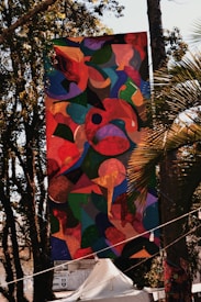 A large, colorful abstract banner is hanging outdoors among trees. The artwork features a mixture of organic shapes in vibrant colors, including reds, greens, blues, and yellows. Sunlight filters through the trees, casting shadows and highlights on the banner. Below the banner, there is a white tent and some signage visible in the background.