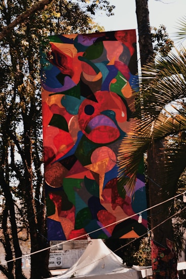 A large, colorful abstract banner is hanging outdoors among trees. The artwork features a mixture of organic shapes in vibrant colors, including reds, greens, blues, and yellows. Sunlight filters through the trees, casting shadows and highlights on the banner. Below the banner, there is a white tent and some signage visible in the background.