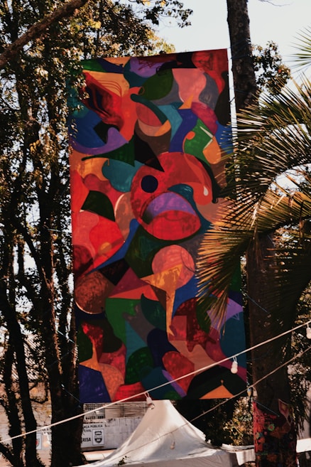 A large, colorful abstract banner is hanging outdoors among trees. The artwork features a mixture of organic shapes in vibrant colors, including reds, greens, blues, and yellows. Sunlight filters through the trees, casting shadows and highlights on the banner. Below the banner, there is a white tent and some signage visible in the background.