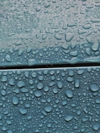 Water droplets of various sizes cover a smooth blue surface, creating a glossy appearance. The droplets are densely packed, emphasizing the wetness contrasting with the matte texture of the surface.