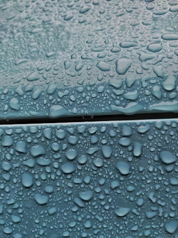 Water droplets of various sizes cover a smooth blue surface, creating a glossy appearance. The droplets are densely packed, emphasizing the wetness contrasting with the matte texture of the surface.