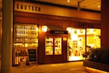 A warmly lit storefront displaying rows of wine bottles, with the word 'ENOTECA' prominently on the facade. There is a wooden door with glass panes flanked by large windows showing the interior where two people are visible, likely engaged in activities related to wine or dining.