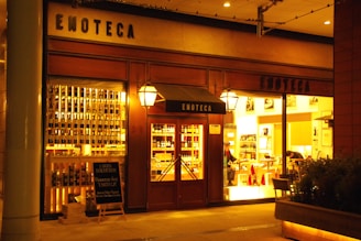A warmly lit storefront displaying rows of wine bottles, with the word 'ENOTECA' prominently on the facade. There is a wooden door with glass panes flanked by large windows showing the interior where two people are visible, likely engaged in activities related to wine or dining.
