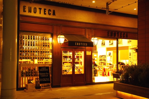 A warmly lit storefront displaying rows of wine bottles, with the word 'ENOTECA' prominently on the facade. There is a wooden door with glass panes flanked by large windows showing the interior where two people are visible, likely engaged in activities related to wine or dining.
