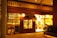 A warmly lit storefront displaying rows of wine bottles, with the word 'ENOTECA' prominently on the facade. There is a wooden door with glass panes flanked by large windows showing the interior where two people are visible, likely engaged in activities related to wine or dining.