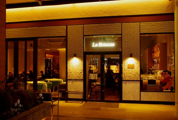 The exterior of a warmly lit restaurant named 'La Brianza' is captured at night. The entrance features glass windows, revealing a cozy, dimly lit interior with tables and seated patrons. A playful cartoon character is visible on one of the window panels. The facade includes textured brick elements and soft ambient lighting.