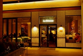 The exterior of a warmly lit restaurant named 'La Brianza' is captured at night. The entrance features glass windows, revealing a cozy, dimly lit interior with tables and seated patrons. A playful cartoon character is visible on one of the window panels. The facade includes textured brick elements and soft ambient lighting.