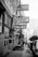 A black and white street scene with vintage signs for services like '4 Hour Cleaning' and 'Valet Cleaners Tailor' hanging above a sidewalk. People are sitting at tables outside, and a few others are walking along the street. The atmosphere is urban and bustling, with various storefronts visible along the street.