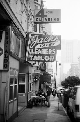 A black and white street scene with vintage signs for services like '4 Hour Cleaning' and 'Valet Cleaners Tailor' hanging above a sidewalk. People are sitting at tables outside, and a few others are walking along the street. The atmosphere is urban and bustling, with various storefronts visible along the street.