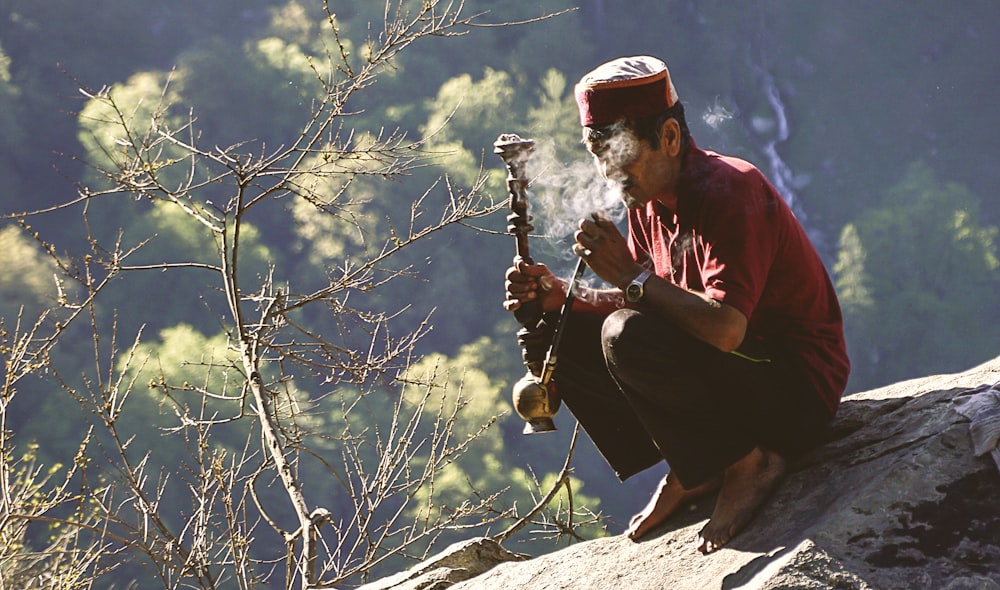 man sitting on rock formation near cliff while holding smoking pipe during daytime