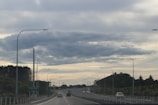 A wide, empty highway stretches into the distance. Street lamps line both sides of the road, and a few scattered vehicles are seen traveling in both directions. Lush green trees and foliage border the highway. Overhead, the sky is filled with soft, diffused clouds, giving a serene environment. A sign reads 'Peka Peka this exit'.