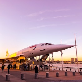 An airplane is displayed outdoors as part of an exhibit, with several people nearby. The setting is at sunset, offering a warm and nostalgic atmosphere. The airplane is elevated on platforms with stairs leading up to it. The backdrop includes water and a skyline of buildings.