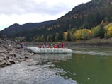 A group of people wearing life jackets are seated on a white inflatable raft labeled 'Fraser River Expeditions,' floating near a rocky riverbank. The river is surrounded by a dense forest with autumn foliage, featuring a mix of green and yellow trees. The background shows forested hills under a cloudy sky.