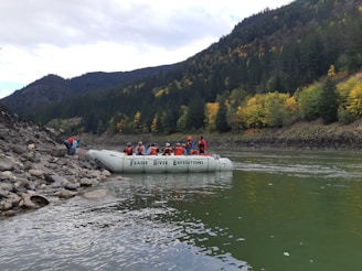 A group of people wearing life jackets are seated on a white inflatable raft labeled 'Fraser River Expeditions,' floating near a rocky riverbank. The river is surrounded by a dense forest with autumn foliage, featuring a mix of green and yellow trees. The background shows forested hills under a cloudy sky.