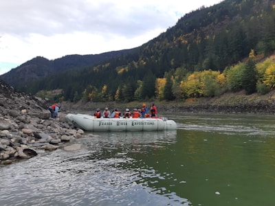 A group of people wearing life jackets are seated on a white inflatable raft labeled 'Fraser River Expeditions,' floating near a rocky riverbank. The river is surrounded by a dense forest with autumn foliage, featuring a mix of green and yellow trees. The background shows forested hills under a cloudy sky.