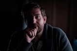 A person with a contemplative expression is sitting in a dimly lit environment, wearing a dark-colored robe or sweater. The light casts shadows on their face, emphasizing the somber atmosphere.