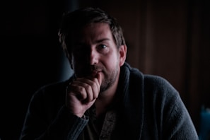A person with a contemplative expression is sitting in a dimly lit environment, wearing a dark-colored robe or sweater. The light casts shadows on their face, emphasizing the somber atmosphere.