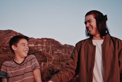 A candid photo of two friends laughing together on a sunlit mountain trail, backpacks on and the horizon stretching wide behind them.