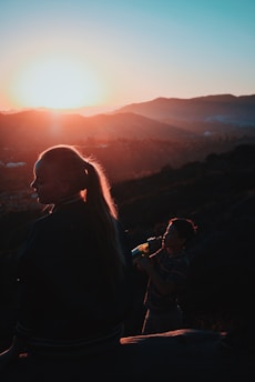 Silhouettes of a person with long hair and a child with a bottle stand against a mountainous landscape during sunset. The sun casts a warm glow across the scene, creating a serene and tranquil atmosphere.