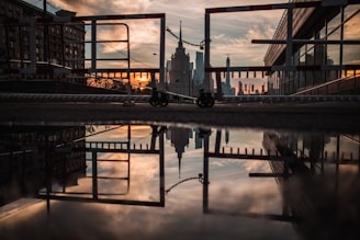 A dramatic sunset is reflected in a puddle on an urban rooftop. The silhouette of city skyscrapers and a series of barriers create a strong contrast against the warm tones of the sky. The symmetry between the reflection and the actual scene adds depth to the composition.