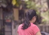 A person with long dark hair tied in a ponytail wearing a pink shirt is facing away. The background is blurred, featuring greenery and out-of-focus structures.