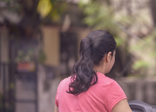 A person with long dark hair tied in a ponytail wearing a pink shirt is facing away. The background is blurred, featuring greenery and out-of-focus structures.