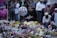 A memorial setup featuring numerous floral tributes and messages. In the foreground, a balloon with the words 'You'll Be Missed' is visible. Several people gather around the site, some holding candles and dressed in diverse attire, suggesting a somber gathering.